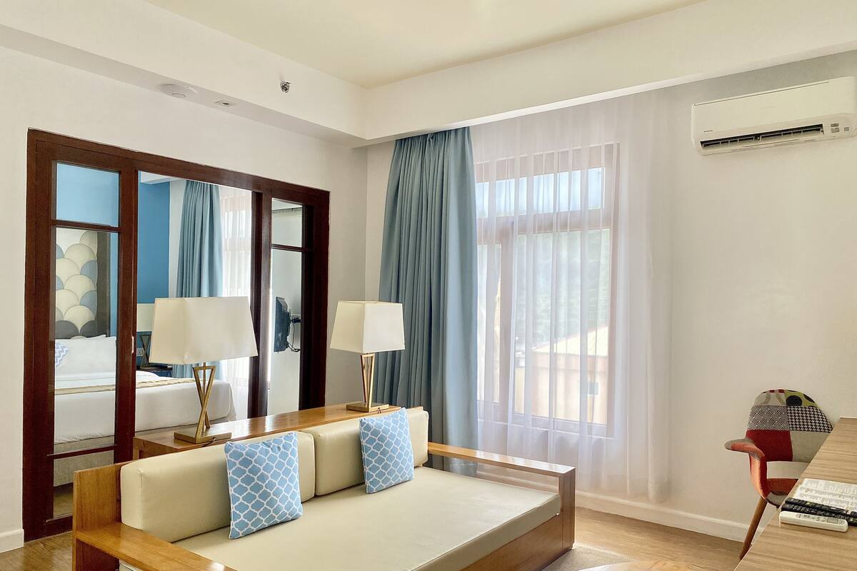 Ein modernes Hotelzimmer mit einer Couch, zwei Lampen, einem Spiegel, einem Fenster mit Vorhängen, einer Klimaanlage und einer Fernbedienung auf einem Holztisch.
