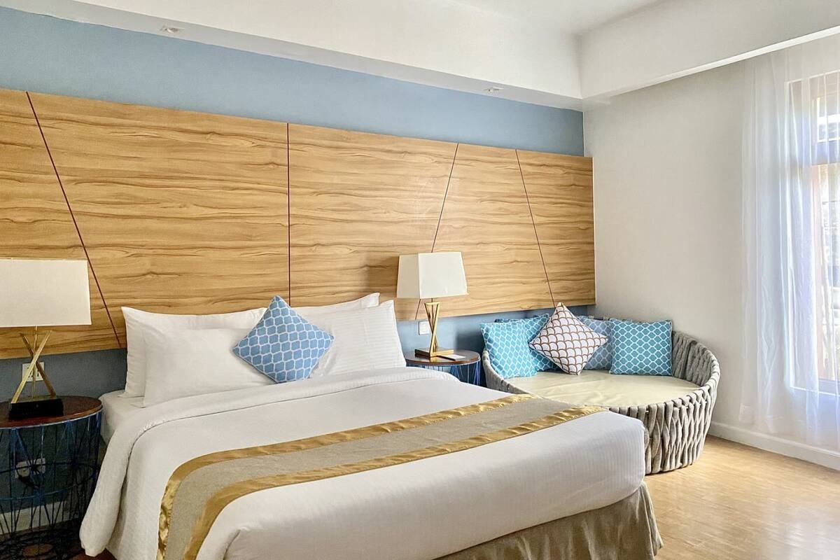 Ein Hotelzimmer mit einem großen Bett, einem Nachttisch, einer Lampe, einem Sofa und einem Fenster mit durchsichtigen Vorhängen.