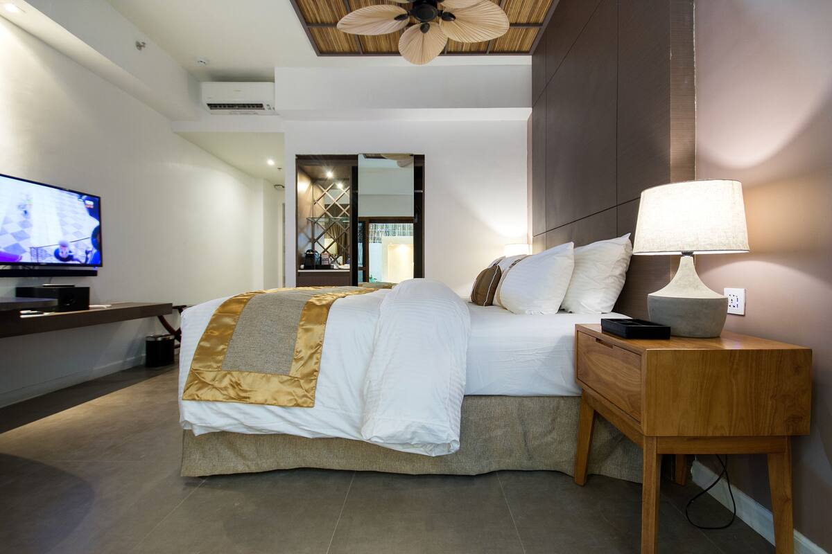 Ein modernes Hotelzimmer mit einem großen Bett, einem Nachttisch mit Lampe, einem Fernseher links und einem Holz-Kopfteil.