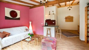 A cozy living room with a white sofa, a small coffee table, and a colorful ottoman. There's a kitchen area with a bar stool and a hanging plant. - Arrive - holidays - refueling in "House on Forest Park" (Scheidegg-Lindenau)