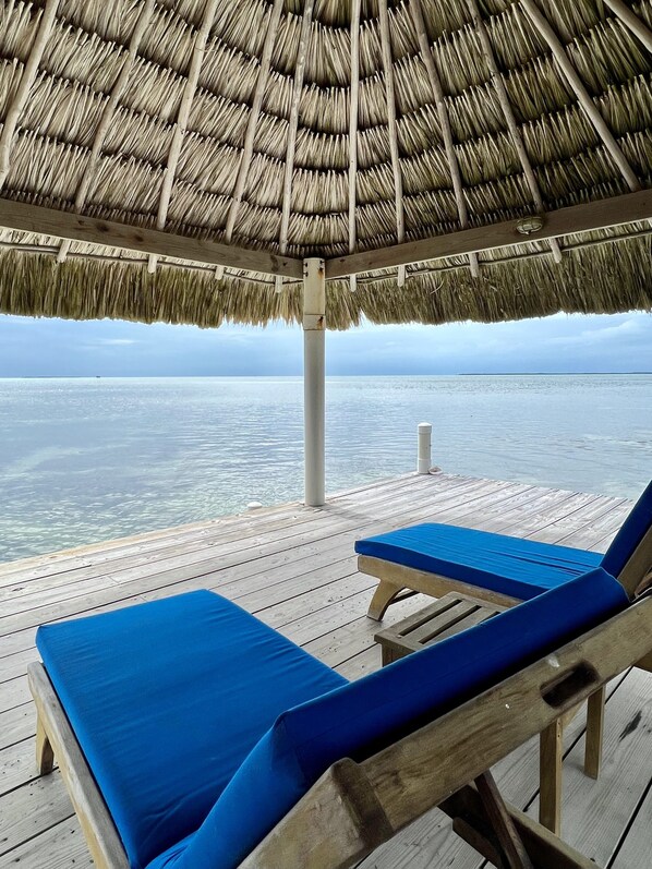 A wooden deck with two blue cushioned lounge chairs under a thatched roof, overlooking a calm sea.