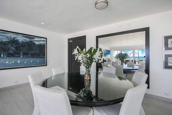A modern dining area with a glass table surrounded by white chairs, a large painting, and a mirror reflecting a room with a view of the sea.