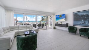 Een moderne woonkamer met een witte modulaire bank, een groene poef en een aan de muur bevestigde televisie. Er is een groot raam met uitzicht op het strand en palmbomen.