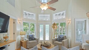 A spacious living room with a high ceiling, large windows, and a ceiling fan.