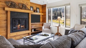 Een gezellige woonkamer met een open haard, houten wanden, een bank, een bijzettafeltje en uitzicht op een besneeerd landschap.