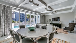 A modern living room with a round dining table, grey upholstered chairs, a ceiling fan, and a large window overlooking a pool and buildings. - Luxury North Beach Resort home w/ pool/elevator & marsh/ocean view (North Myrtle Beach)