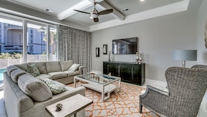 A modern living room with a sofa, coffee table, and armchair, a glass-top table, a dark cabinet, and a large window with a view of buildings. - Luxury North Beach Resort home w/ pool/elevator & marsh/ocean view (North Myrtle Beach)