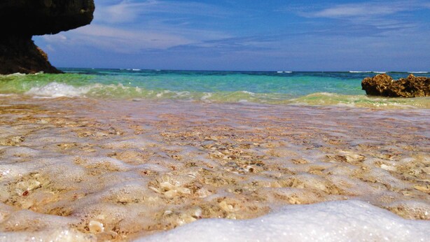 A beach with clear shallow waters, visible rocks, and a large rock formation in the background. - Private Lodge COTENCHI (Nakijin)