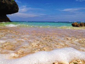 A beach with clear shallow waters, visible rocks, and a large rock formation in the background.