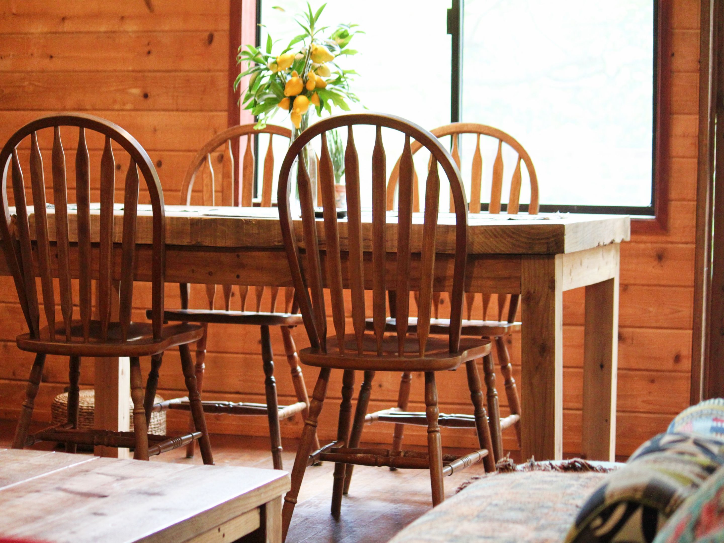 Una mesa de comedor de madera con cuatro sillas, un florero con flores amarillas y una ventana al fondo.
