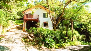 A rustic house with a wooden balcony, surrounded by lush greenery and a stone pathway. - Private Lodge COTENCHI (Nakijin)