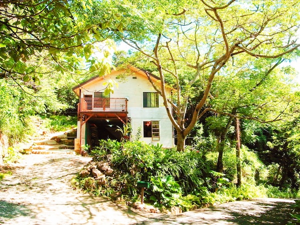A rustic house with a wooden balcony, surrounded by lush greenery and a stone pathway.