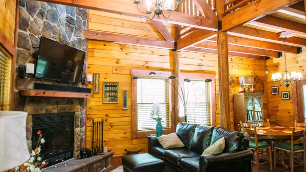 A log cabin interior with a stone fireplace, leather sofa, dining table, and wooden beams.