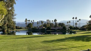 A golf course with a pond, palm trees, and mountains in the background.