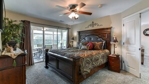 A bedroom with a large bed, a ceiling fan, a nightstand, a lamp, a dresser, and a window with curtains.