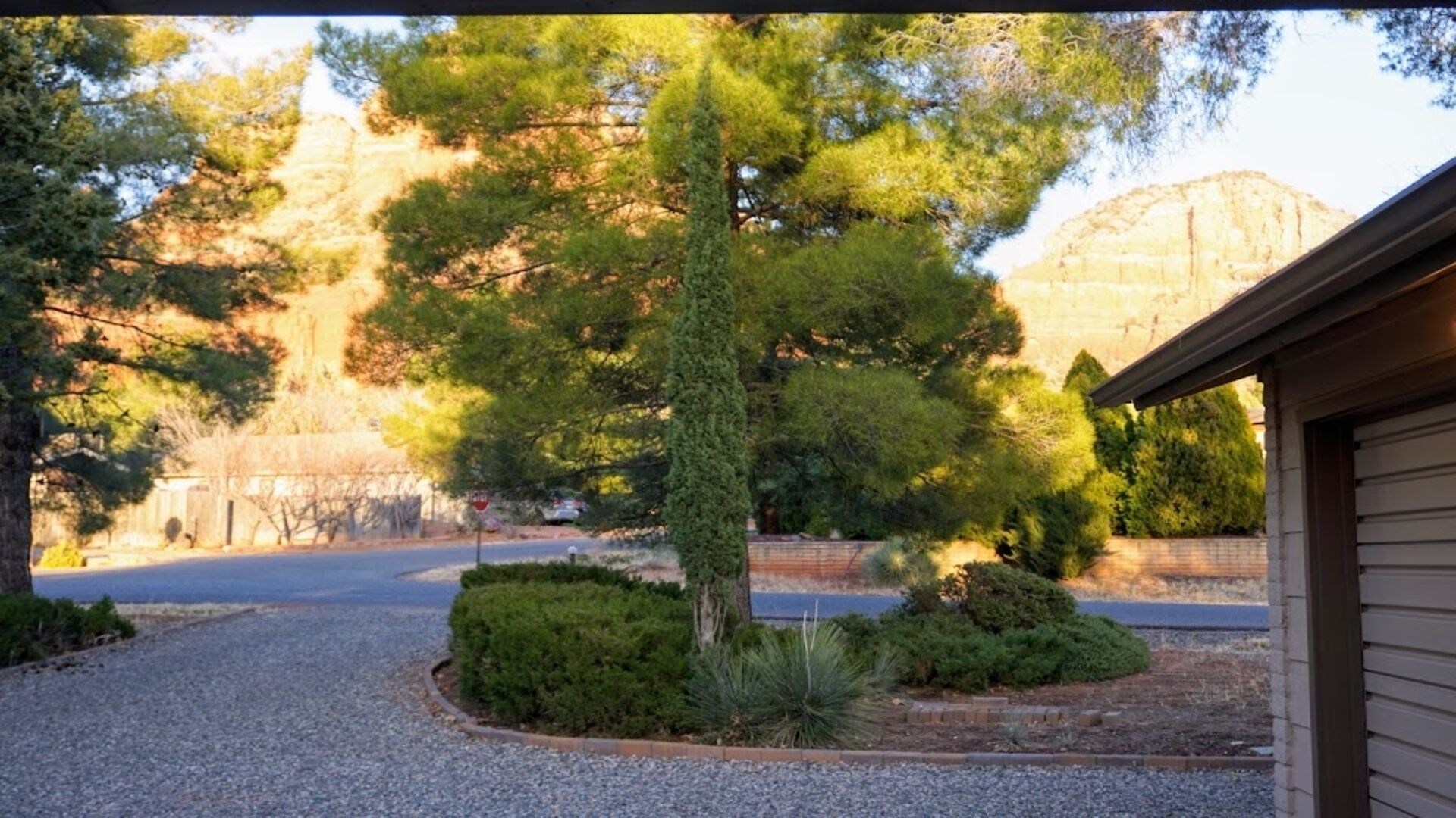 Un carrer d'accés a una vivenda amb un jardí ben cuidat, un arbre conífer alt i un paisatge de muntanya de fons.
