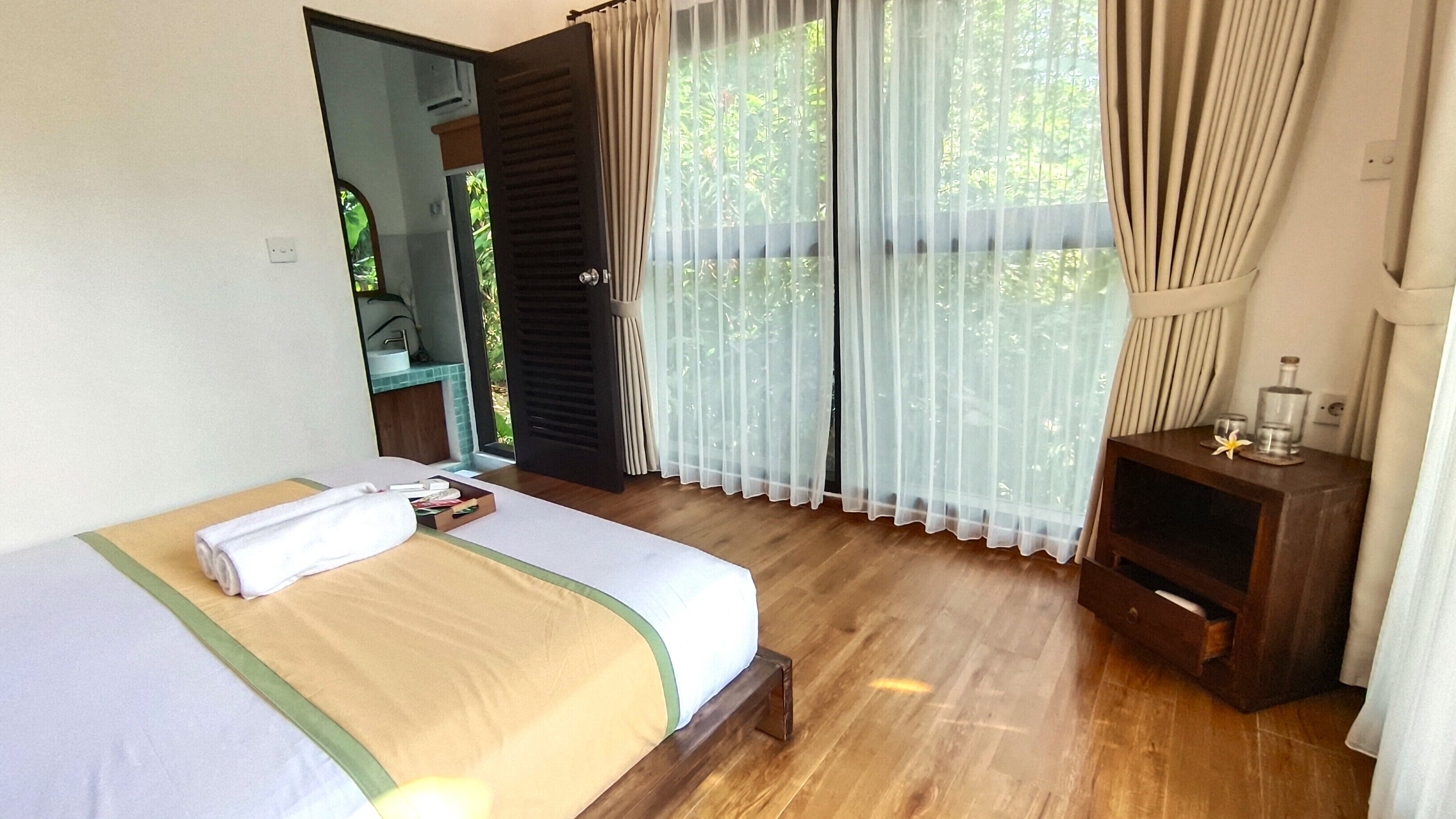 A hotel room with a bed, a wooden nightstand, a mirror, and a window with sheer curtains.