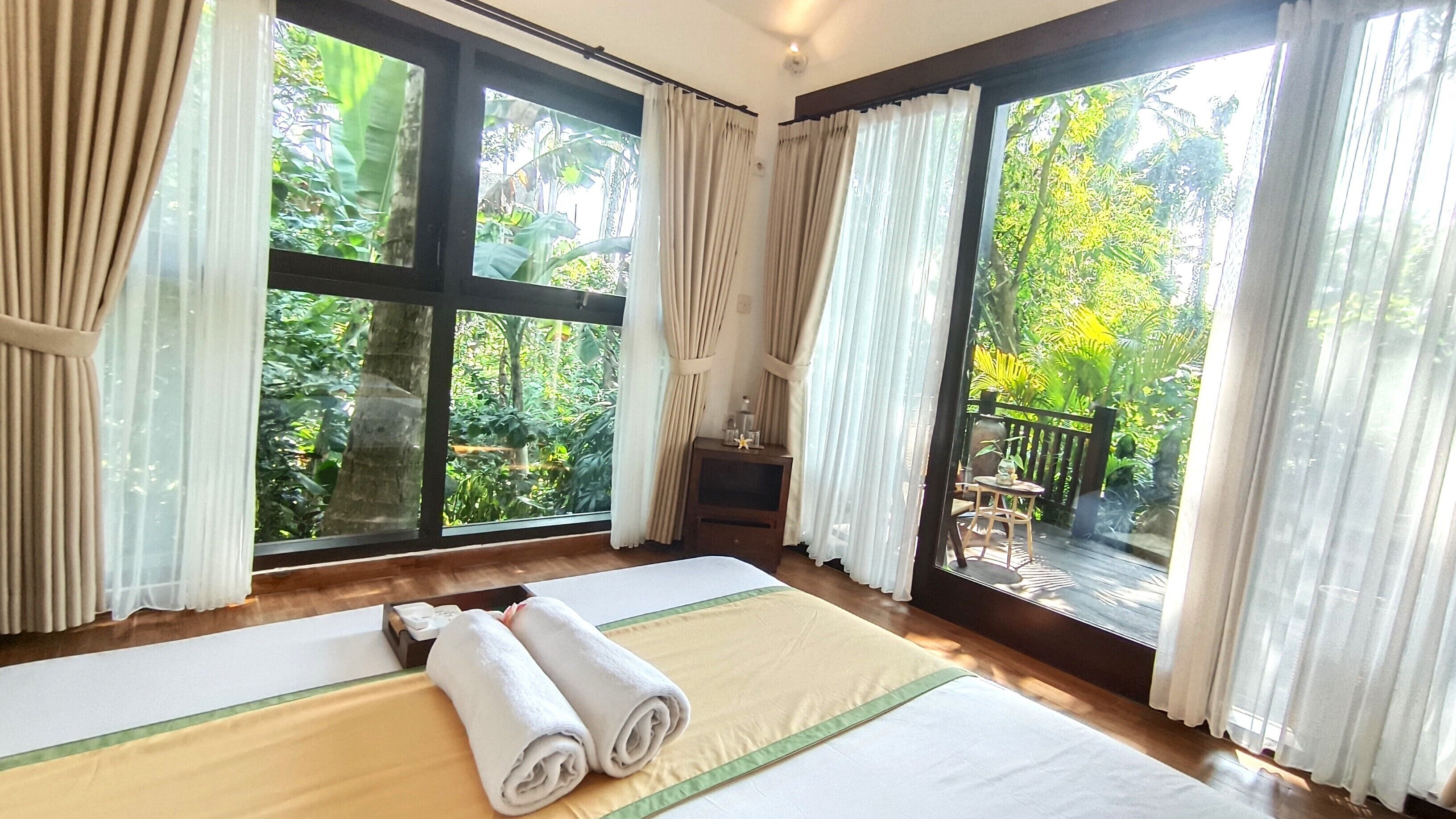 A room with a large bed, a wooden floor, and a view of a lush garden through large windows.