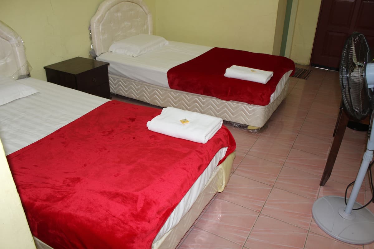 Habitación de hotel con dos camas, cada una con colcha roja y ropa blanca. Hay una mesita de noche entre las camas y un ventilador de pie en la esquina.