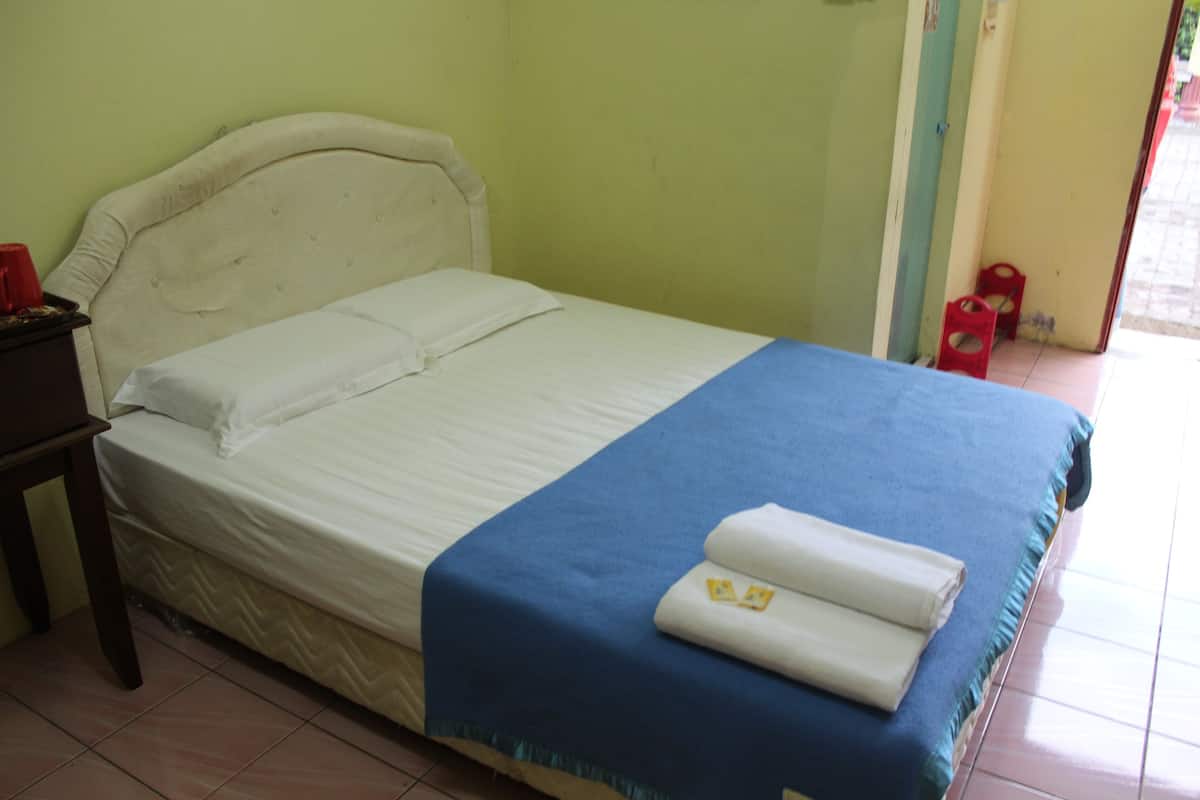 Una cama con cabecera blanca y colcha azul, una taza roja sobre una mesita, y una puerta con un taburete rojo afuera.