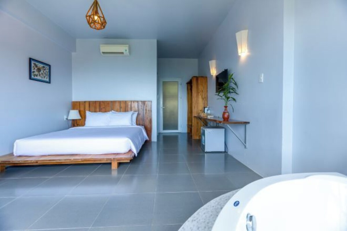 A modern hotel room with a large bed, a small desk, a chair, a potted plant, and a bathtub.