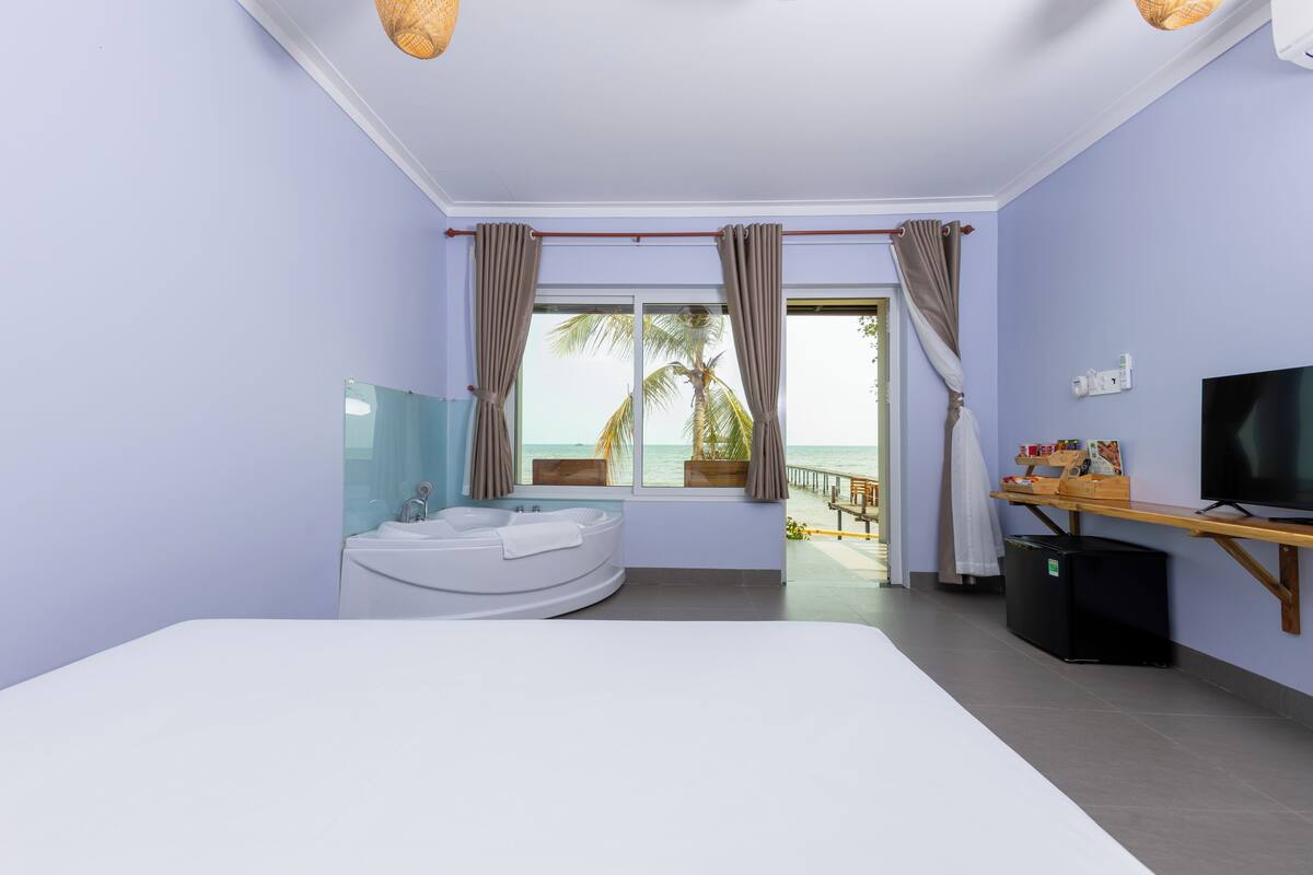 A bedroom with a large bed, a bathtub, a TV, and a view of the beach.