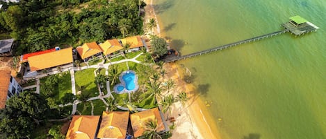 An aerial view of a resort with a swimming pool, beach, and a pier extending into the water.