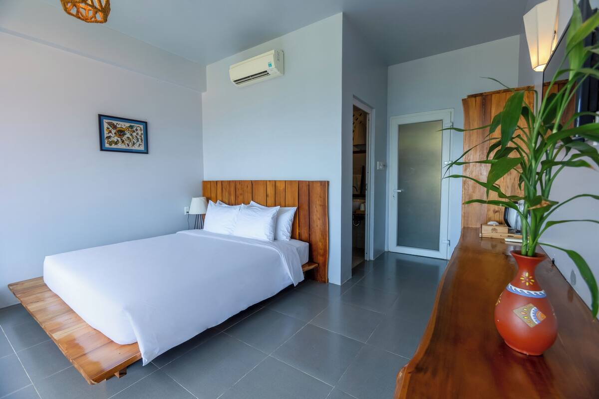 A modern hotel room with a wooden headboard, a white bed, a red vase with a plant, and a wooden bench.