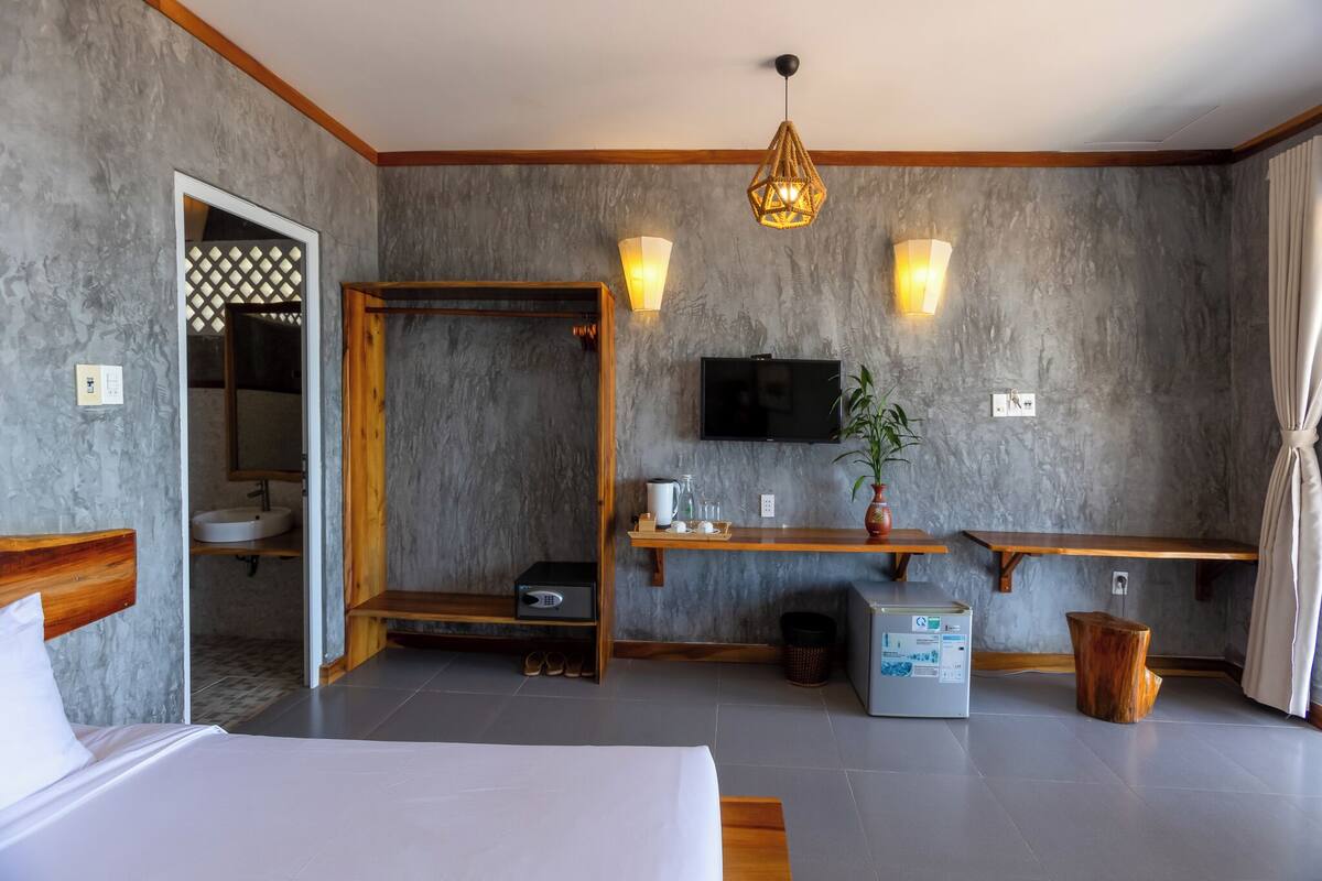 A modern hotel room with a bed, a TV, a wooden bench, a small table with a plant, and a bathroom visible through an open door.