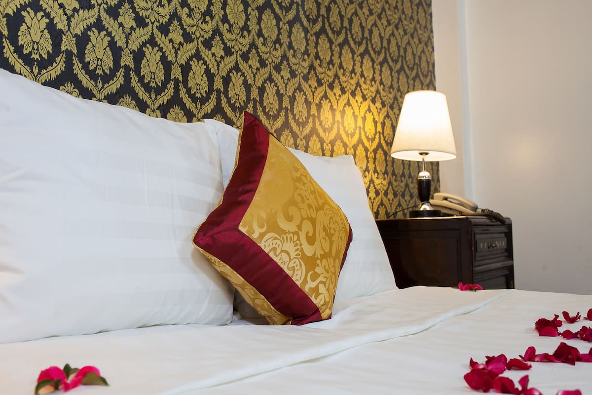 Un lit parfaitement fait, avec des draps blancs, un coussin rouge et doré, et des pétales de rose éparpillés sur le lit.
