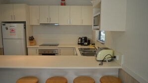 A kitchen with white cabinets, a stainless steel refrigerator, a stove, a toaster, and three wooden stools. - Discobulous 6 (Mount Buller)