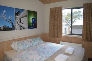 Un dormitorio con cabecera de madera, cama con colcha estampada, dos almohadas, ventana con cortinas y cuadros con escenas de la naturaleza.