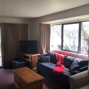 A living room with a television on a wooden stand, a sofa with a red blanket, a wooden coffee table, and a window with curtains. - Discobulous 6 (Mount Buller)