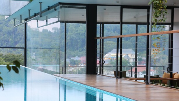 Una piscina interior moderna con paredes de vidrio transparente, suelo de madera y vistas a un área boscosa.
