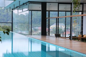 A modern indoor swimming pool with clear glass walls, wooden flooring, and a view of a forested area. - Lakewood Residence (Kandy)