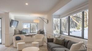 Un salon moderne avec un canapé gris, des fauteuils blancs et une table basse. Un téléviseur est fixé au mur, et une grande fenêtre offre une vue sur les arbres et la neige à l’extérieur.
