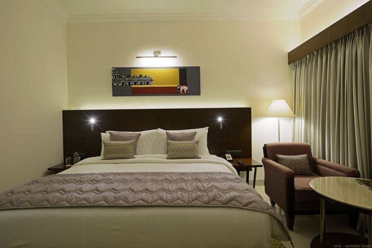 A hotel room with a large bed, a chair, a small table, a lamp, and a wall-mounted artwork.