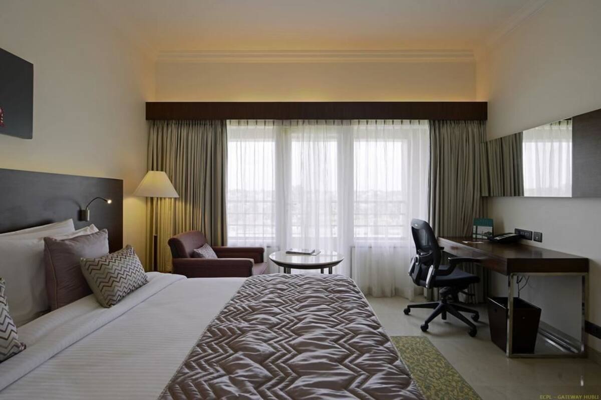 A hotel room with a large bed, a desk, a chair, a lamp, and a view of the city through large windows.