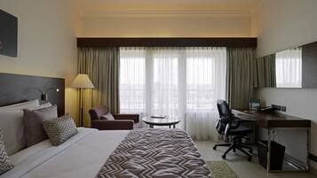 A hotel room with a large bed, a desk, a chair, a lamp, and a view of the city through large windows.
