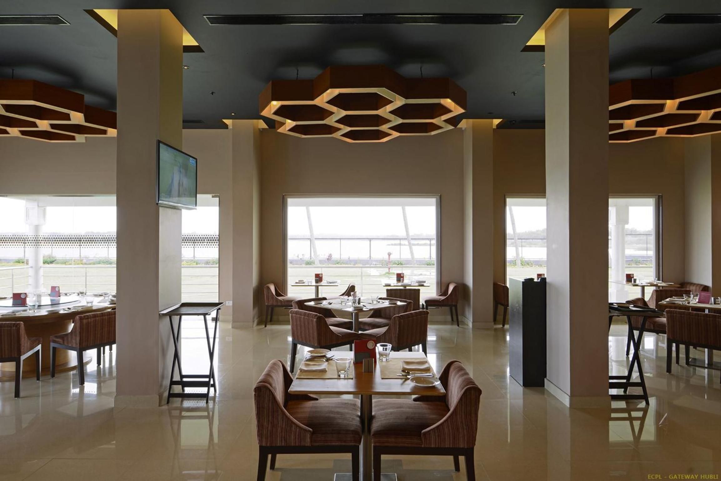 A spacious hotel lobby with high ceilings, modern hexagonal light fixtures, and large windows offering views of the airport.