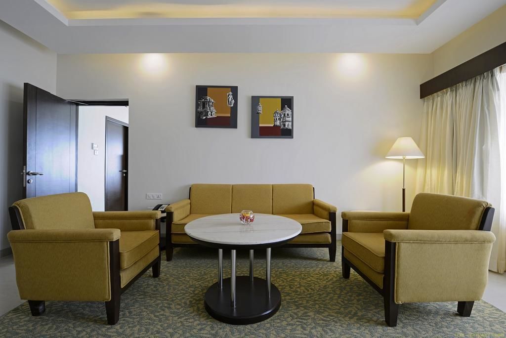 Habitación de hotel con sofá amarillo, dos sillones, una mesa redonda y dos obras de arte colgadas en la pared.
