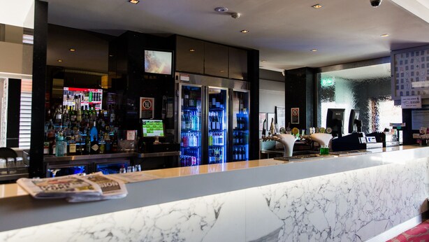 A hotel lobby with a marble reception desk, a bar area with various bottles, and a large screen displaying information. - Nags Head Hotel (Adamstown)