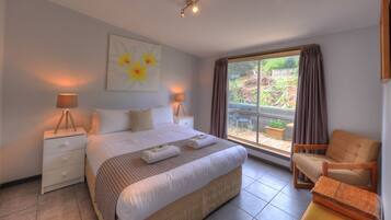 A bedroom with a large bed, two bedside lamps, a nightstand, a chair, and a view of the garden through a sliding glass door.