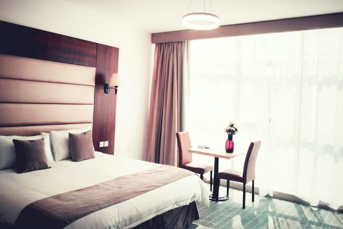 A hotel room with a large bed, a desk, and a chair, a window with curtains, and a hanging light fixture.