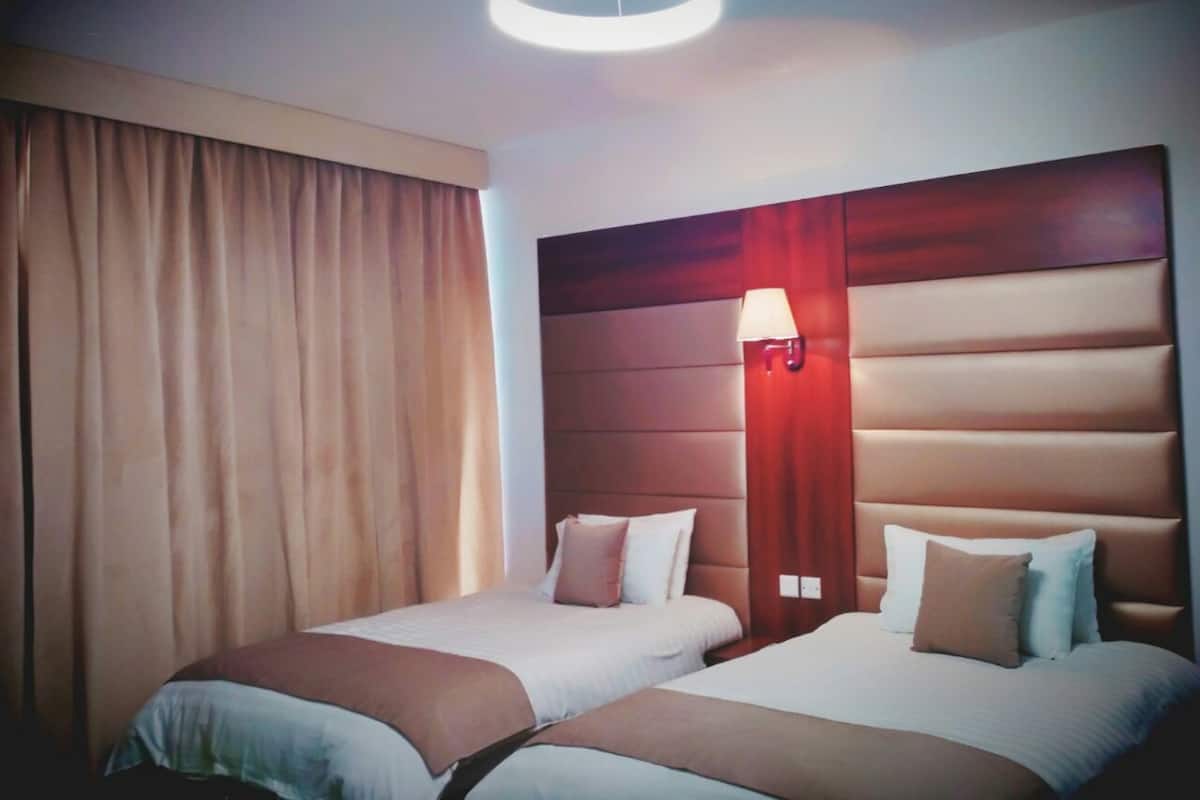 A hotel room with two beds, a large window with curtains, a modern headboard, and a circular light fixture.