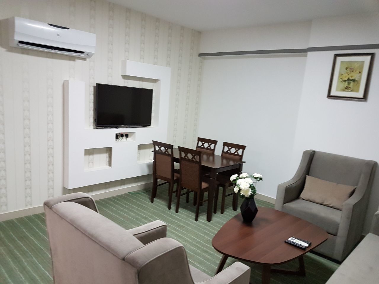 Un salon comprenant un canapé, une table basse avec un vase, une table à manger avec des chaises, un téléviseur à écran plat fixé au mur et un appareil de climatisation.