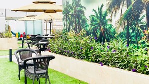 Un espacio al aire libre con sillas de mimbre y sombrillas, un mural de palmeras y un jardín con flores púrpuras.