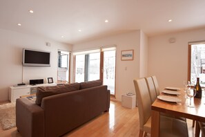 A modern living room with a brown sofa, a flat-screen TV, and a dining area with a wooden table set for two. - Avalon (Kutchan)