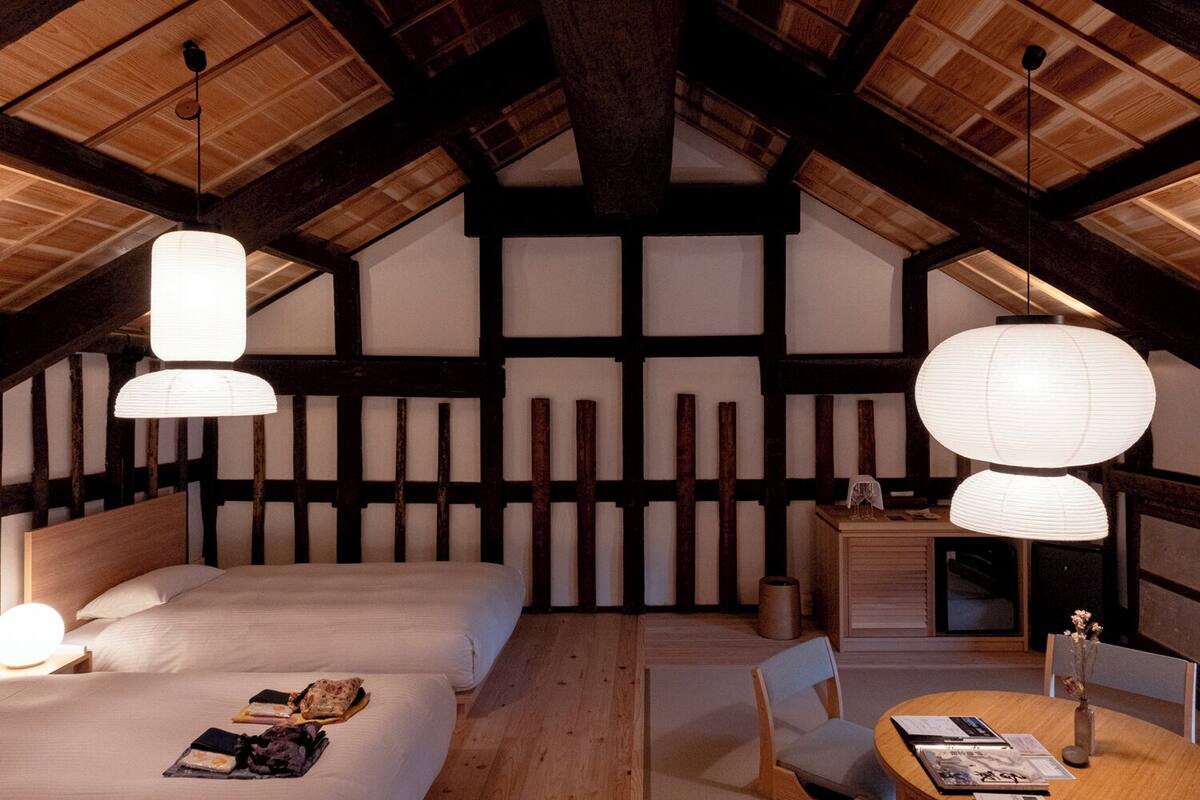 Ein gemütliches Zimmer mit hohen Decken, freiliegenden Holzbalken, einem Bett mit Kissen und Buch, einem kleinen Tisch mit Vase und Speisekarte sowie einer Hängelampe.