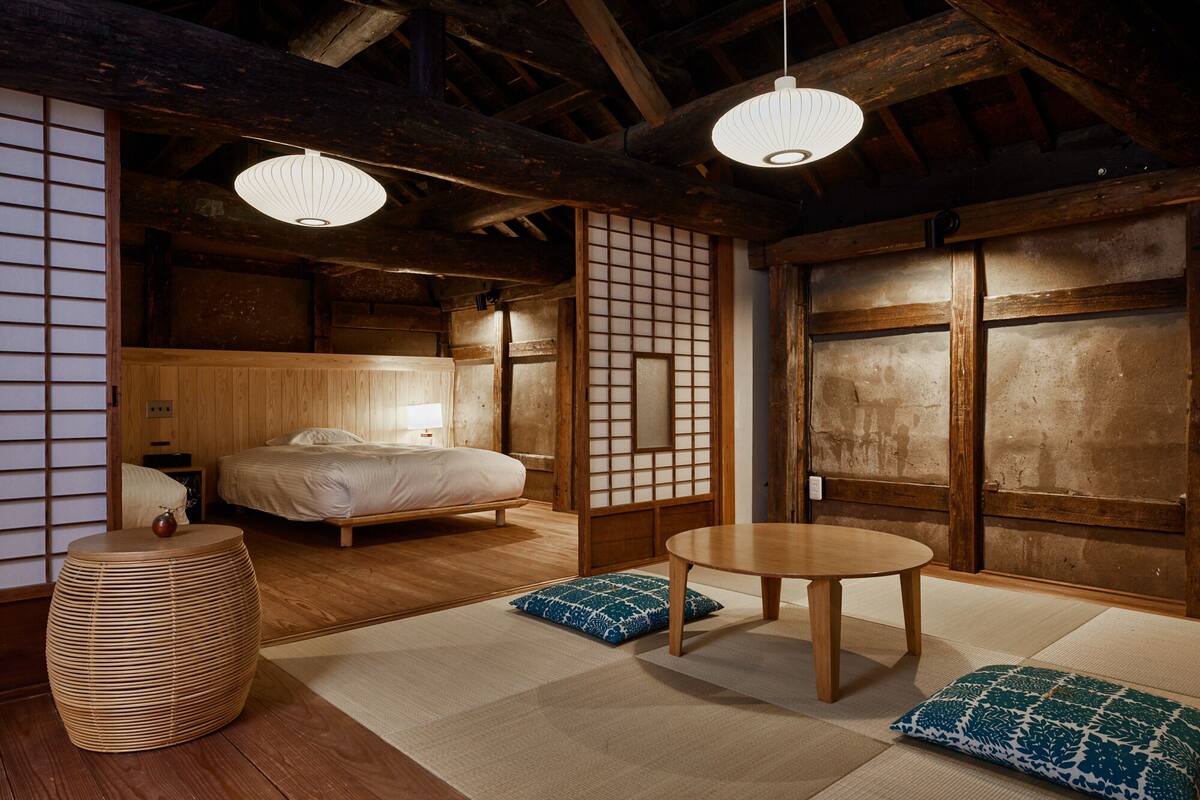Ein traditioneller japanischer Raum mit Tatami-Boden, einem niedrigen hölzernen Bett, einem runden Holztisch und zwei hängenden Papierlaternen.